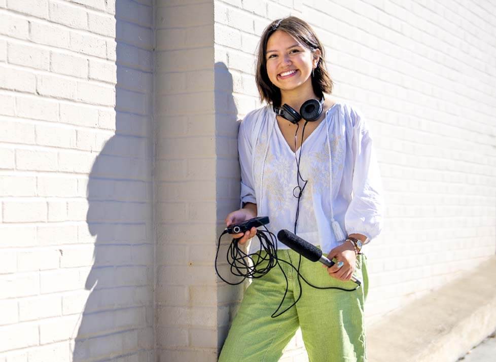 A photo of a student standing outside holding a microphone and headphones